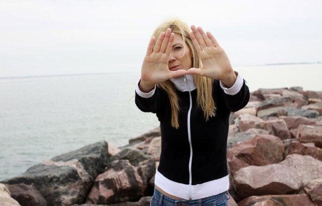 A woman stands outdoors in nature, holding her hands up in front of her face with her thumbs and index fingers forming a rectangle, like a film director framing a shot. She is looking through the space between her hands, focusing on the view.