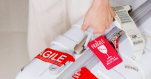 Nha Trang, Vietnam - August 16, 2016: Man holding suitcase in hand at airport of Cam Ranh. Bright red luggage tag (Fragile, Handle with care) attached to white plastic suitcase.
