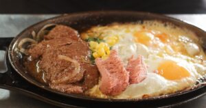 Indulge in the delicious Bò Né Bít Tết, a sizzling Vietnamese steak served with tender meat, a fried egg, fresh vegetables, and crispy bread – a hearty and flavorful meal.