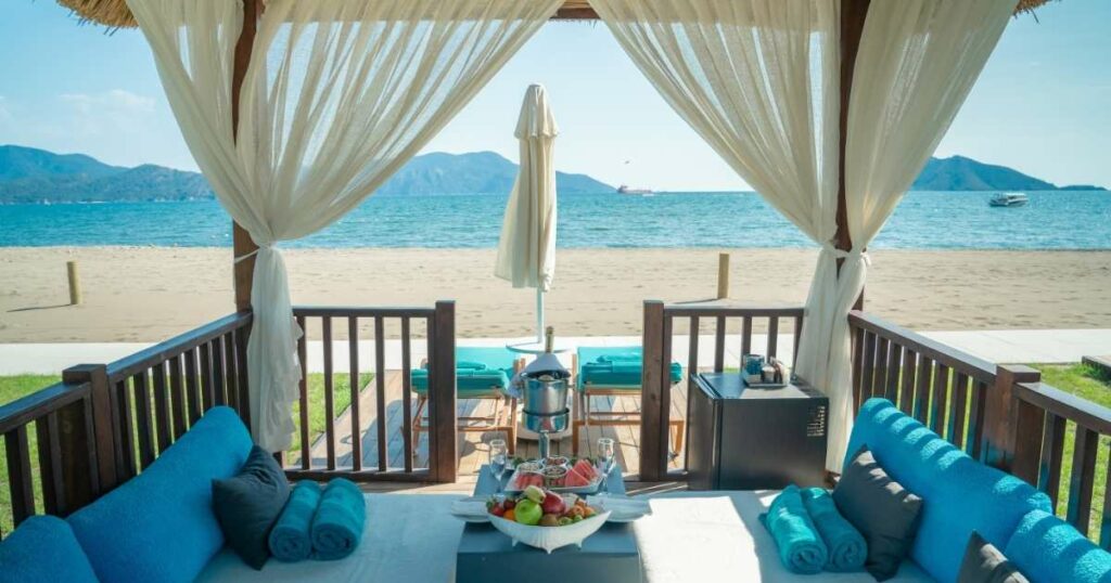 Shutterstock Beautiful beachside boho setup at sunset featuring a bamboo canopy with flowing white curtains, low wooden tables, cozy floor seating with leather poufs, and decorative pampas grass arrangements.