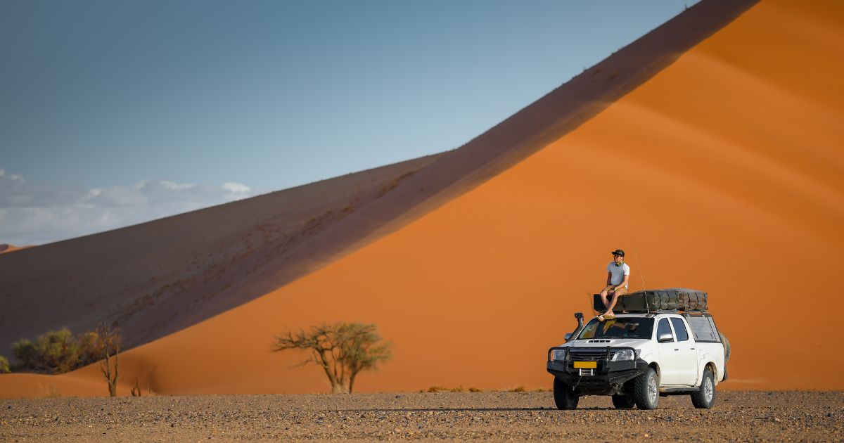 Young Asian man tourist sitting on camper car near orange sand dune landscape. Travel desert in Namibia, Africa.