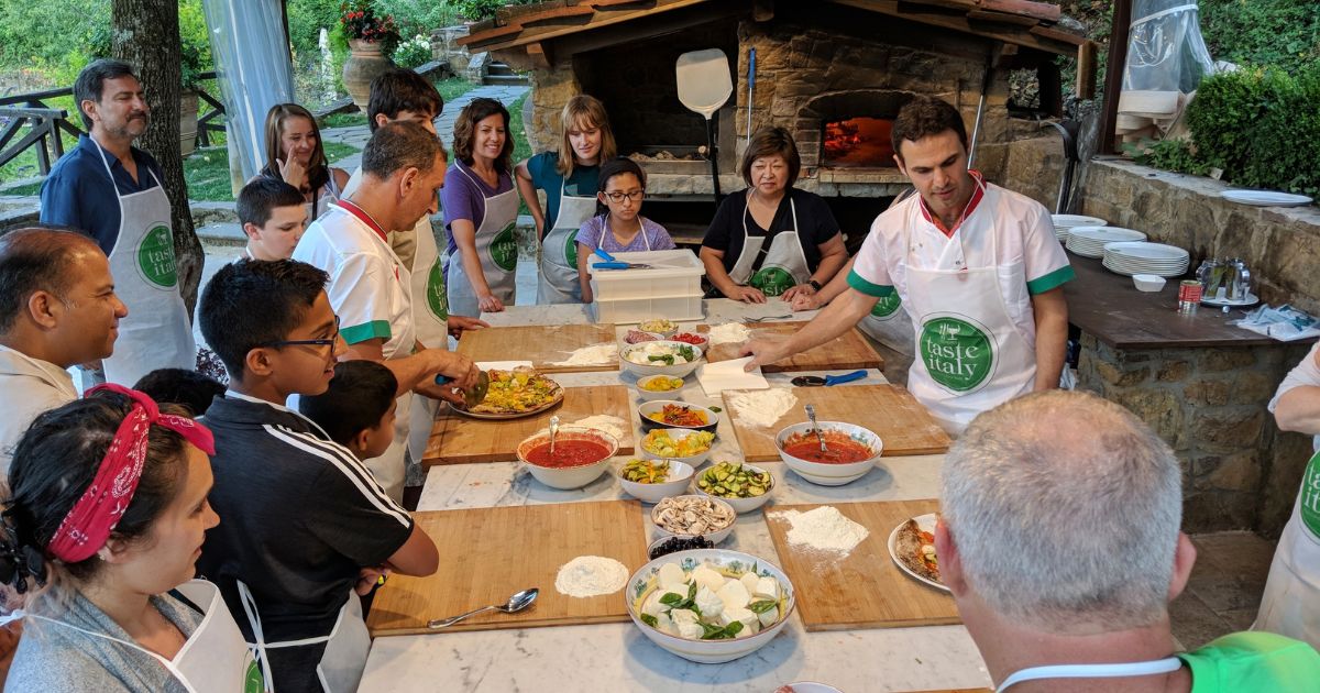 Florence, Tuscany, Italy. June 22, 2018. Cookery classes at Taste Italy. Students and instructors sit and stand at a table with ingredients for Italian pizza. A wood burning oven is in the background,