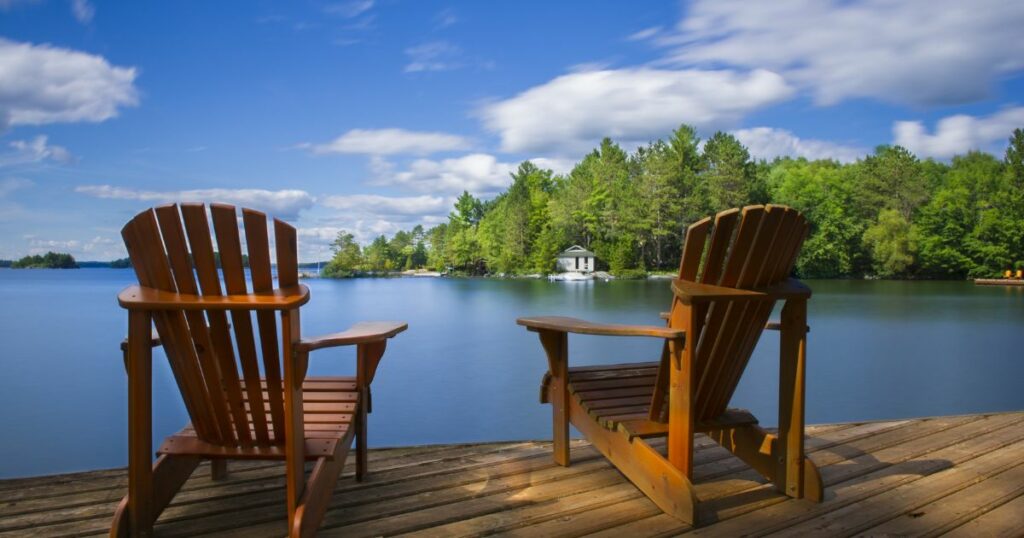 Shutterstock Two Muskoka chairs sitting on a wood dock facing a calm lake. Across the water is a white cottage nestled among green trees. There is a boat dock on the water in front of the cottage.