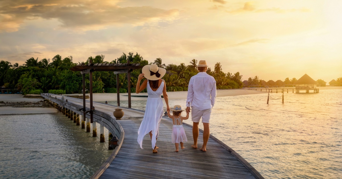 A family in white summer clothes walks holding hands over a wooden pier towards a tropical island in the Maldives during sunset time