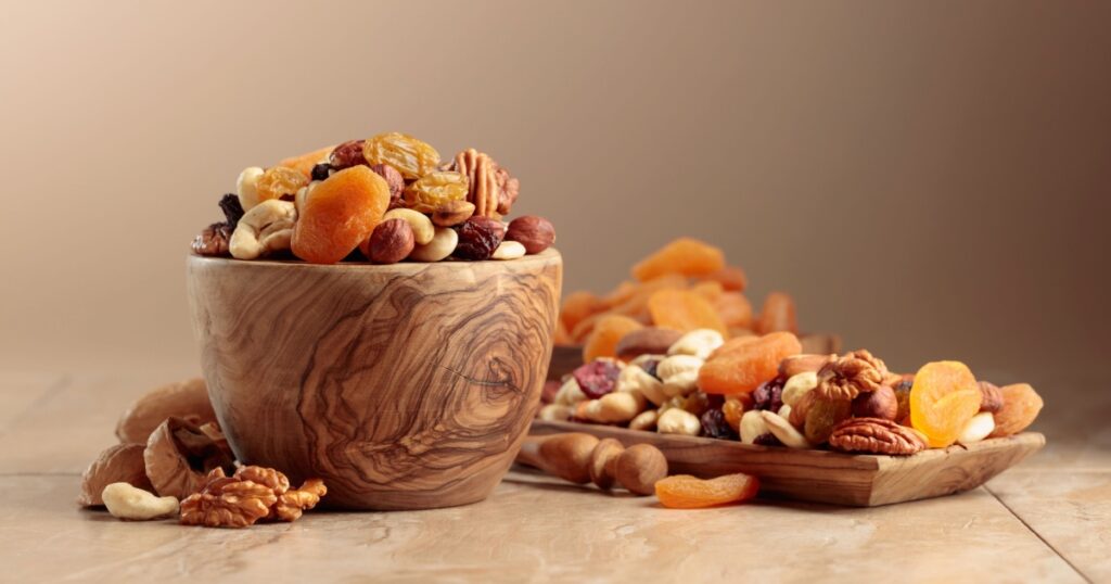 Dried fruits and nuts on a beige ceramic table. The mix of nuts, apricots, and raisins in a wooden bowl.