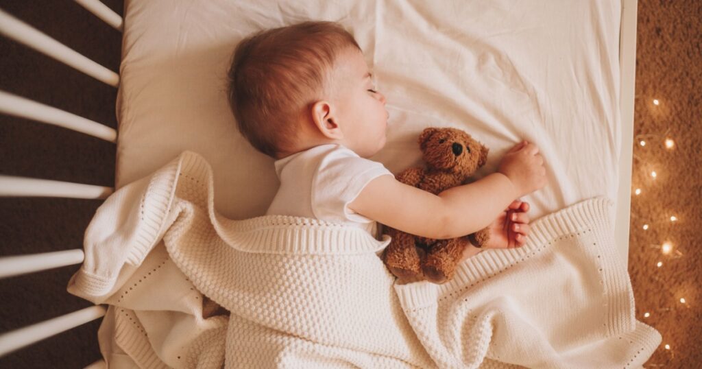 A little baby in a white bodysuit sleeping in a white crib holding a teddy bear and surrounded by small lights