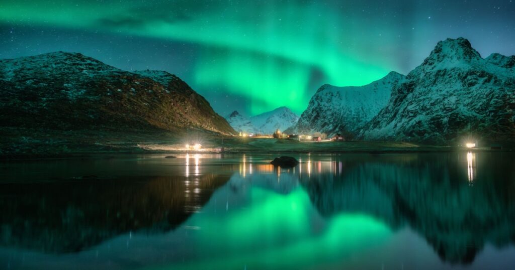 Aurora borealis, snowy mountains, sea, fjord, reflection in water, street lights at starry winter night. Lofoten, Norway