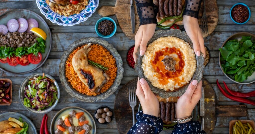 Hands of two women holding tradiitional meal harries or keşkek at the dining table with many meals.