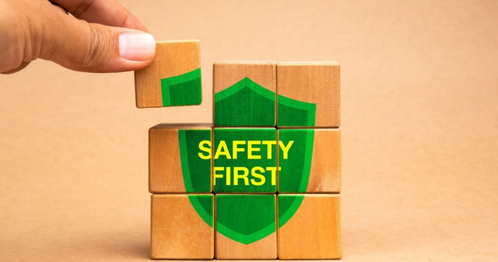 Hand holding a last piece, put on the wooden cube block puzzle stack with text "SAFETY FIRST" on green shield icon isolated on brown background.