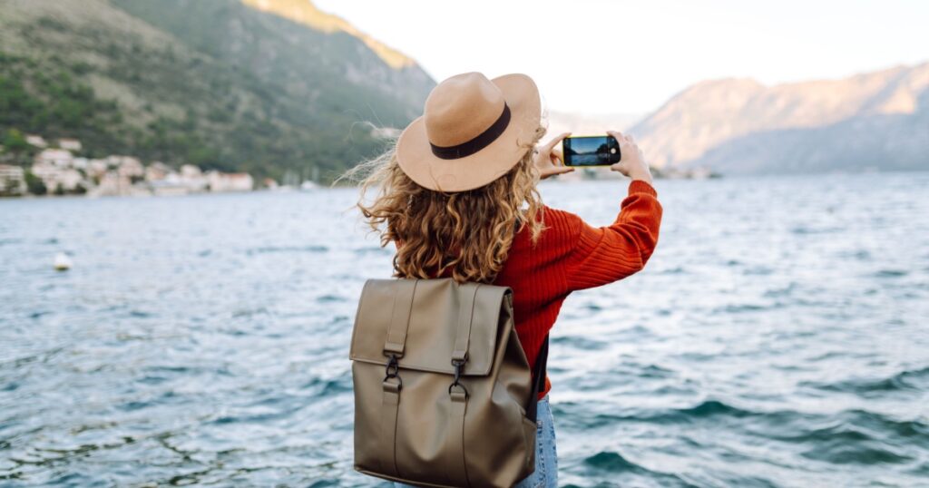 Female traveler in casual sweater taking picture of amazing landscape with mountains, ocean and sunset sky on smartphone during vacation