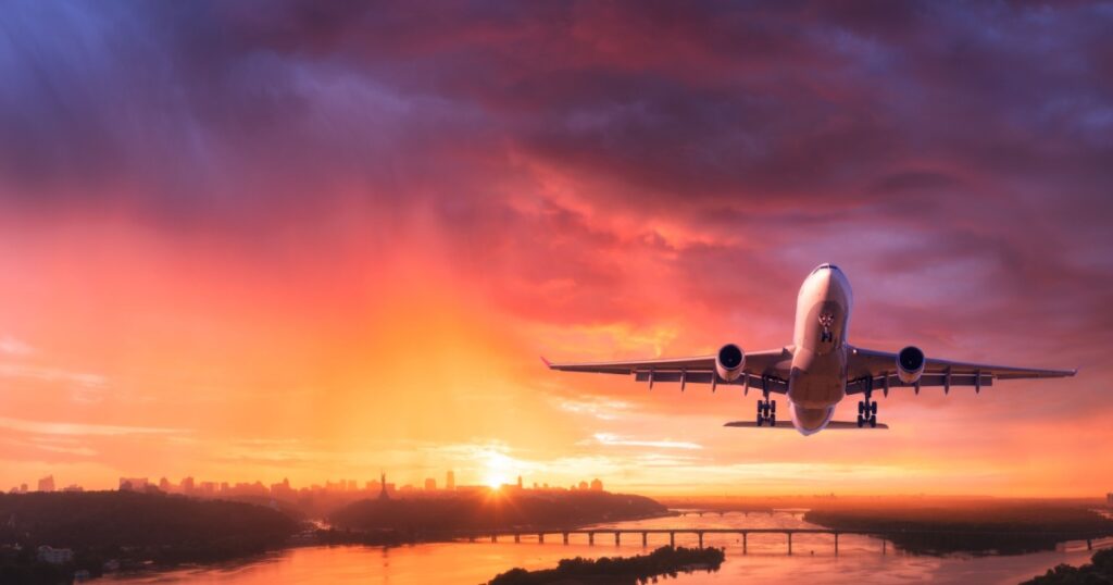 Airplane is flying in colourful sky over city at sunset.