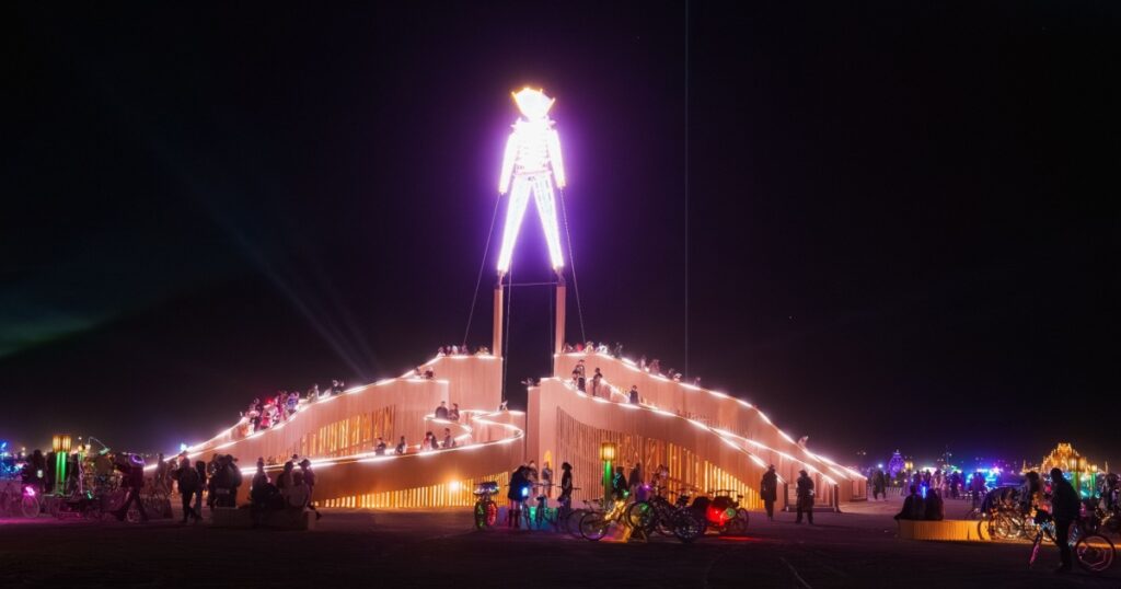 A large, glowing Man stands on a tiered structure at night, surrounded by participants and bicycles with colorful lights at Burning Man in Nevada.