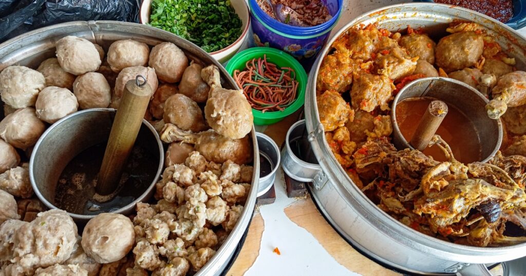 A traditional Indonesian bakso (meatball) food stall setup, featuring various types of meatballs, chicken, condiments like fried shallots, chopped herbs, noodles, and spicy sambal.