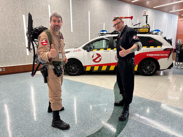 Two cosplayers dressed as Ghostbusters, complete with proton packs, pose with a Ghostbusters-themed vehicle at Fan Expo Canada.