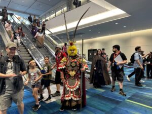 A cosplayer in a detailed golden and red warrior costume with tall headpieces stands proudly while people admire their outfit.