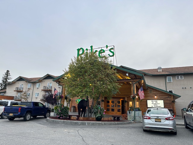 Front entrance of Pike’s Waterfront Lodge in Fairbanks, Alaska, with log cabin–style exterior, American flags, and large green neon sign reading “Pike’s.” Cars are parked outside under an overcast evening sky.