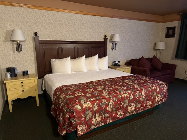 Guest room at Pike’s Waterfront Lodge with a king-size bed covered in a floral quilt, white pillows, wooden headboard, side tables with lamps, and a sofa in the corner.