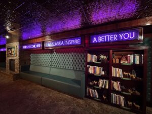 Cozy neon-lit lounge area at Pike’s Waterfront Lodge with plush seating, a bookshelf filled with titles, and glowing purple signs reading “Let Alaska Inspire a Better You.”