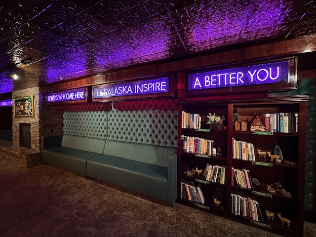 Cozy neon-lit lounge area at Pike’s Waterfront Lodge with plush seating, a bookshelf filled with titles, and glowing purple signs reading “Let Alaska Inspire a Better You.”