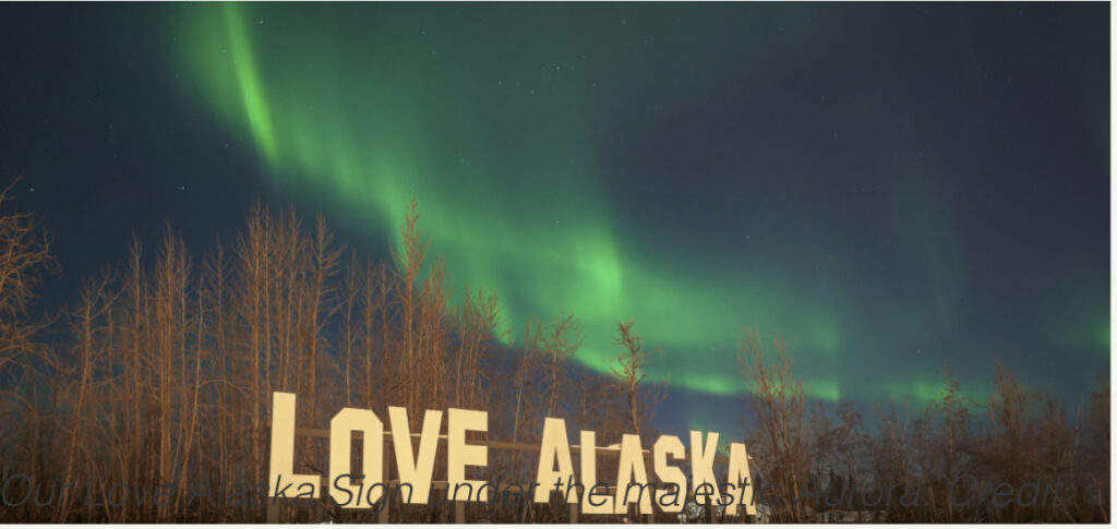 Large outdoor “LOVE ALASKA” sign in bold white letters set against a backdrop of trees and reflected in a small pond at Pike’s Waterfront Lodge with the aurora borealis high above.