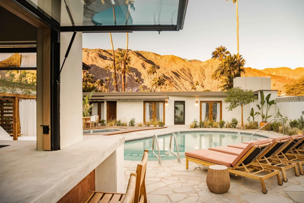 A stylish desert retreat in Palm Springs with a circular swimming pool surrounded by cushioned lounge chairs, palm trees, and modern adobe-style guest rooms, set against the backdrop of golden sunlit mountains.