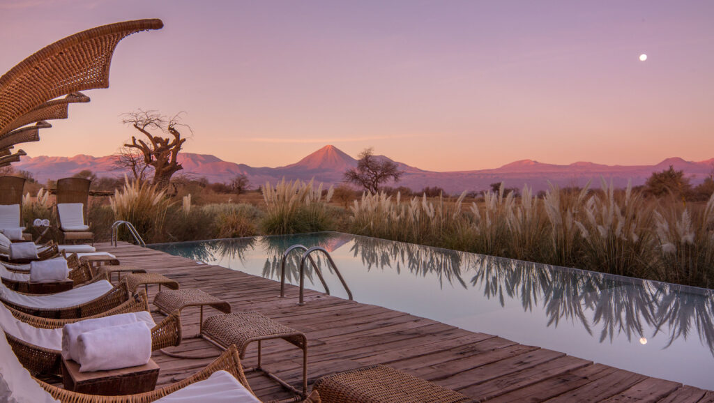A serene infinity pool lined with wicker lounge chairs and white towels overlooks the Atacama Desert at sunset, with pampas grass, a lone tree, and the cone-shaped Licancabur Volcano glowing pink in the distance under a purple sky with the moon rising.