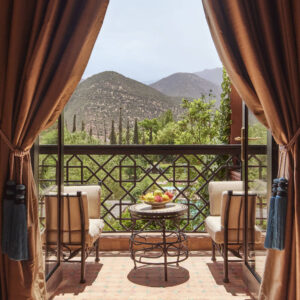 A balcony view framed by draped curtains, with two cushioned chairs and a small table of fruit overlooking lush greenery and distant mountains.
