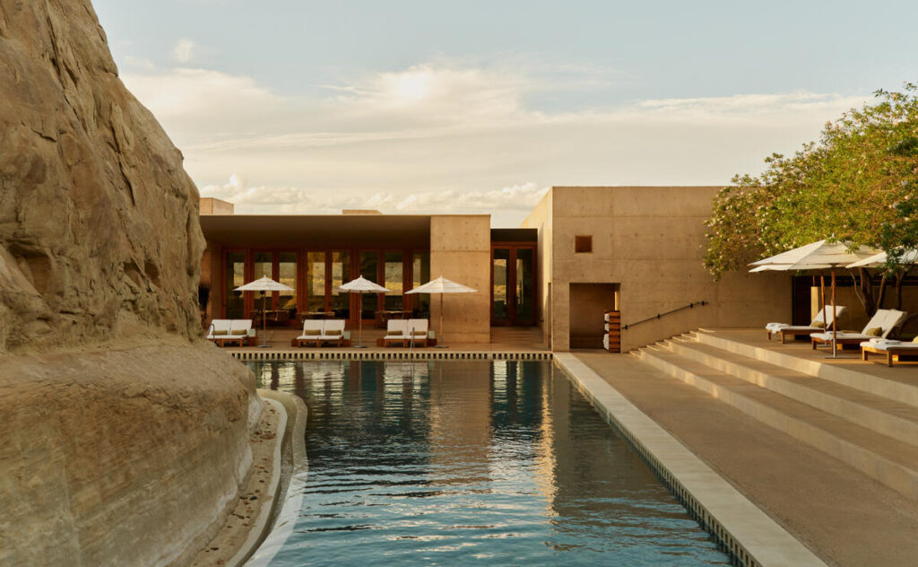 A luxury desert resort featuring a modern stone building with wooden accents, white sun loungers, and umbrellas lining a serene swimming pool beside sandstone cliffs.