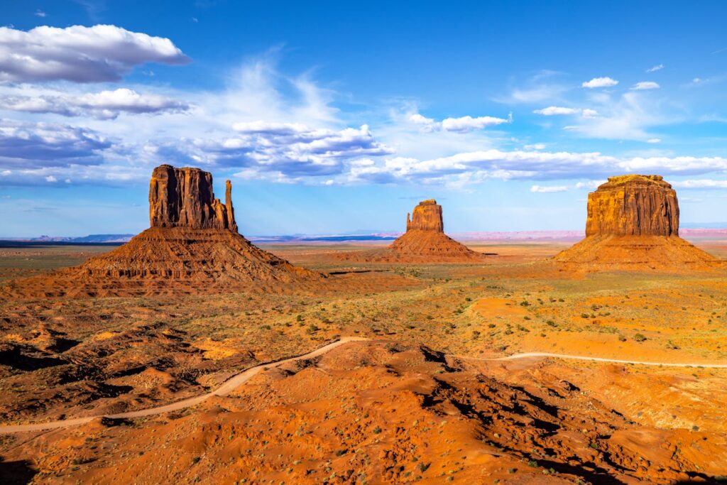 The iconic red sandstone buttes of Monument Valley rise dramatically from the desert floor, surrounded by vast open land under a brilliant blue sky with scattered clouds.