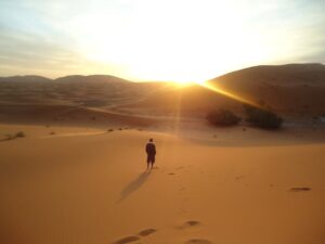 A lone figure walks across golden sand dunes at sunset, leaving footprints behind, as the sun dips low and casts long shadows over the desert hills.