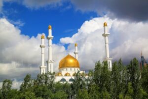 The white Hazrat Sultan Mosque in Astana with golden domes and tall minarets framed by green trees under a dramatic cloudy sky.