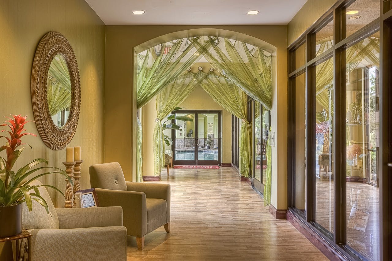 Elegant spa or hotel hallway with soft green curtains draped over an archway, beige armchairs, a decorative mirror, and large windows letting in natural light.