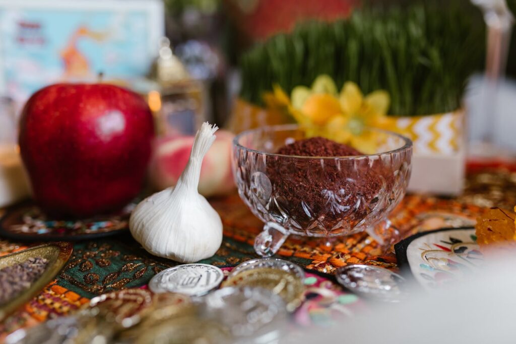 A festive Nowruz table arrangement featuring a crystal bowl of spices, garlic, a red apple, sprouting greenery, flowers, and silver coins.