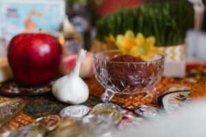 A festive Nowruz table arrangement featuring a crystal bowl of spices, garlic, a red apple, sprouting greenery, flowers, and silver coins.