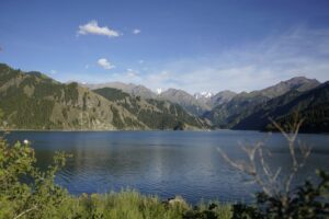 A tranquil alpine lake surrounded by steep green mountains with snow-capped peaks in the distance under a bright blue sky.