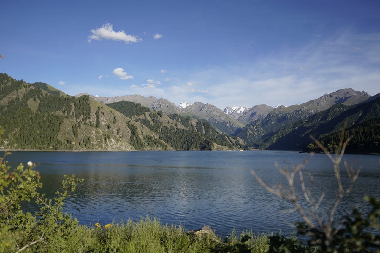 A tranquil alpine lake surrounded by steep green mountains with snow-capped peaks in the distance under a bright blue sky.