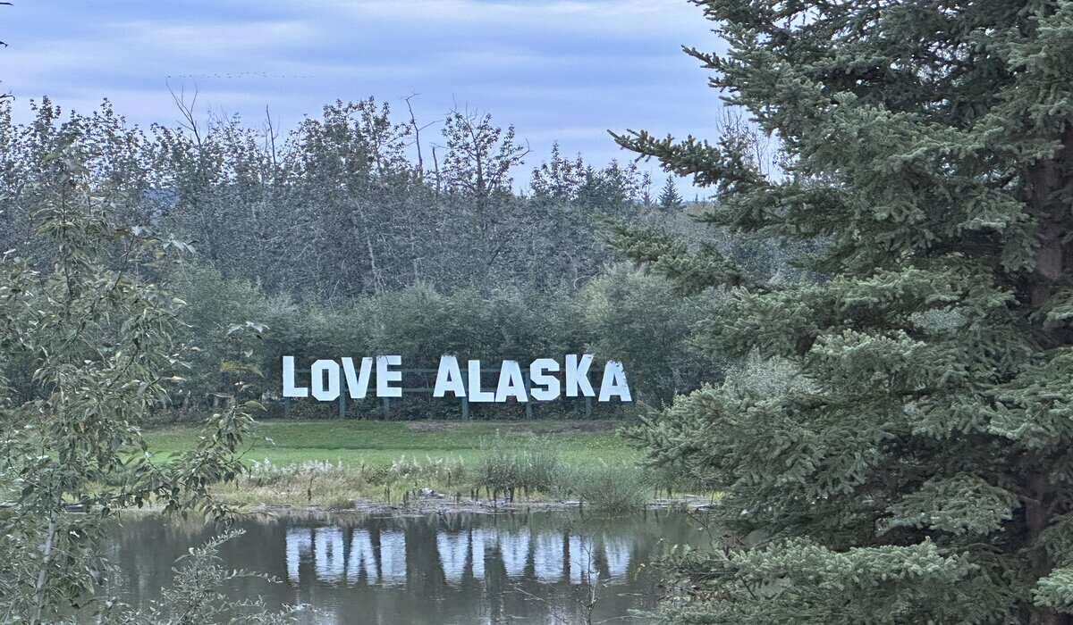 Large outdoor “LOVE ALASKA” sign in bold white letters set against a backdrop of trees and reflected in a small pond at Pike’s Waterfront Lodge.