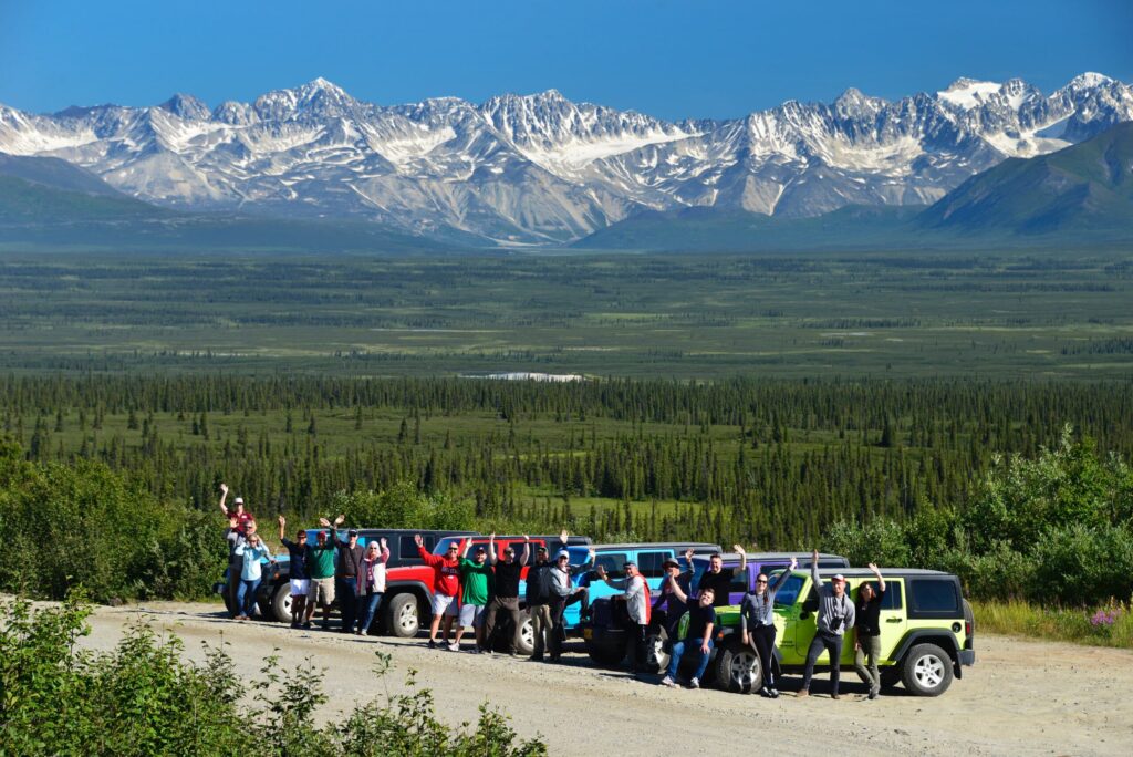Row of colourful Jeep 4x4s with travellers waving beside a pullout; vast forested valley and snow-streaked Alaska Range under blue sky.