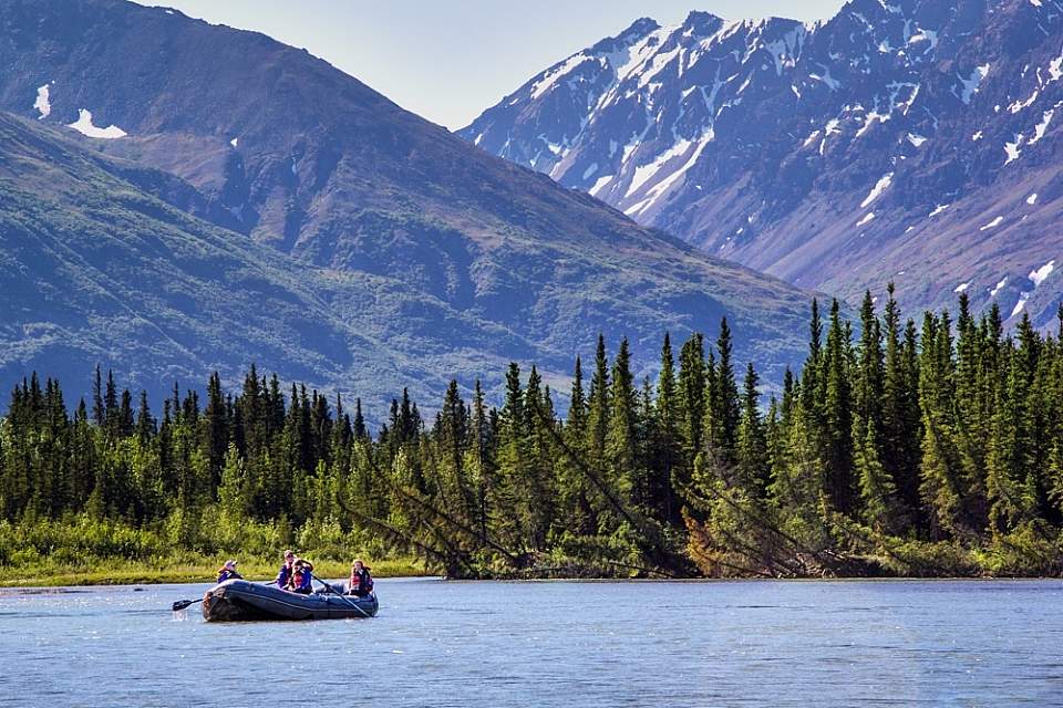 Rafters in a blue raft float a mountain river bordered by dense spruce with rocky peaks behind.
