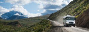 Green Denali transit bus driving a gravel mountain road with a fresh-snow peak ahead.