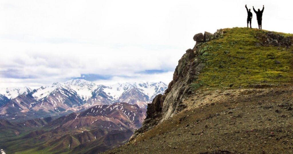 Two hikers silhouetted on a grassy ridge above a panorama of snow-dusted mountains.