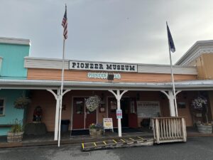 Front entrance of the Pioneer Museum at Pioneer Park in Fairbanks, Alaska, with hanging flower baskets, wooden barrels, and U.S. and Alaska flags flying above.