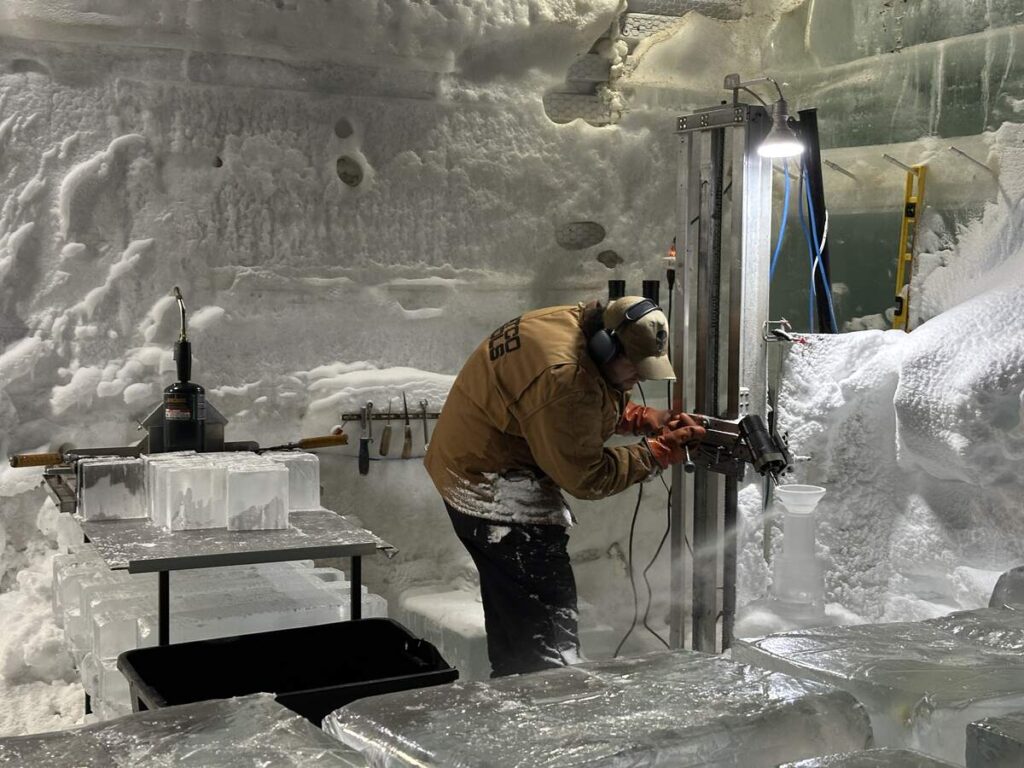Steve Brice carves a martini glass from ice inside the Aurora Ice Museum workshop, surrounded by ice blocks and tools.