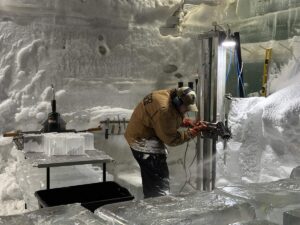 Steve Brice carves a martini glass from ice inside the Aurora Ice Museum workshop, surrounded by ice blocks and tools.