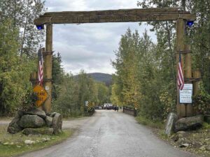 Wooden archway entrance to Chena Hot Springs Resort with American flags, flowers, and a gravel road leading inside.