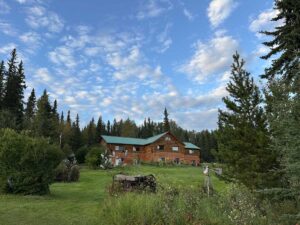 A log lodge with a green roof sits on manicured lawns under a partly cloudy blue sky, framed by trees and garden sculptures.