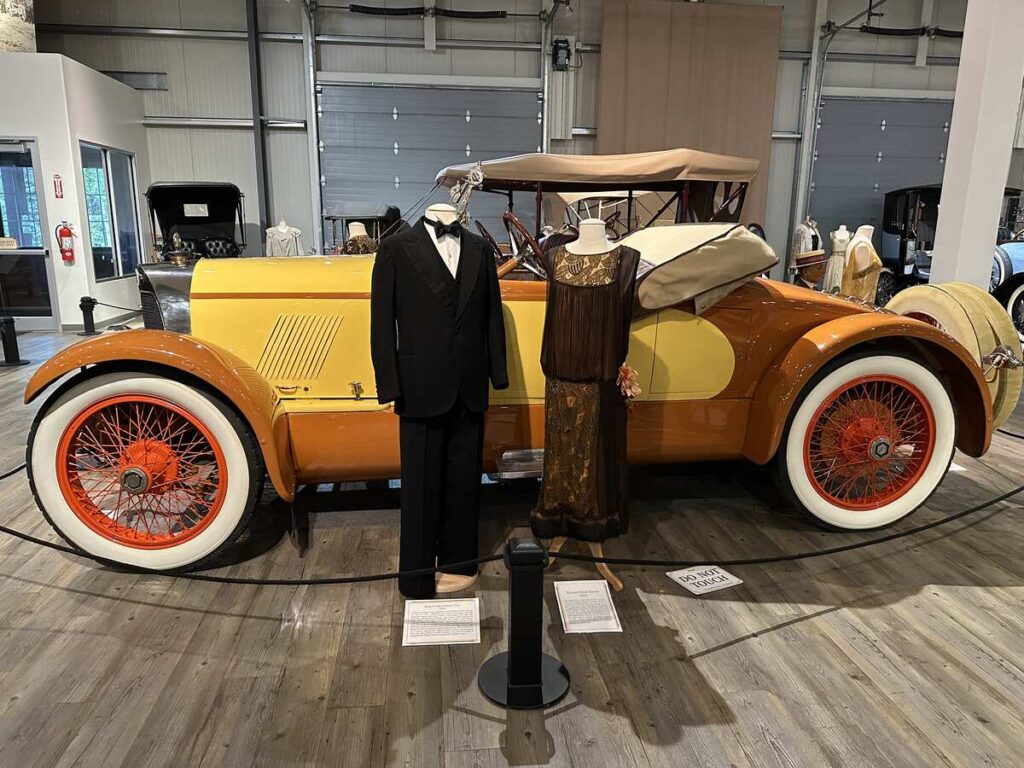 A gold and orange vintage automobile displayed with a tuxedo and flapper-era dress at the Fountainhead Antique Auto Museum.