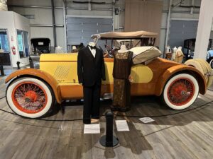 A gold and orange vintage automobile displayed with a tuxedo and flapper-era dress at the Fountainhead Antique Auto Museum.