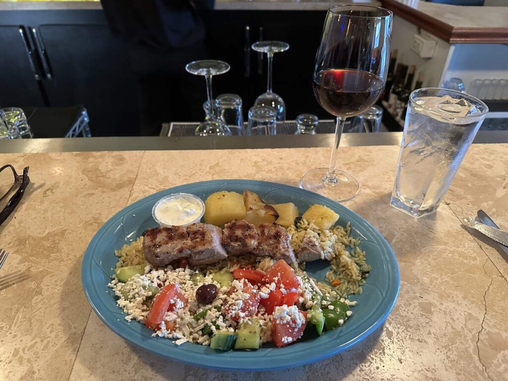 A plate of pork souvlaki with roasted potatoes, rice, Greek salad, and tzatziki, served with a glass of red wine at Bobby’s Restaurant in Fairbanks.