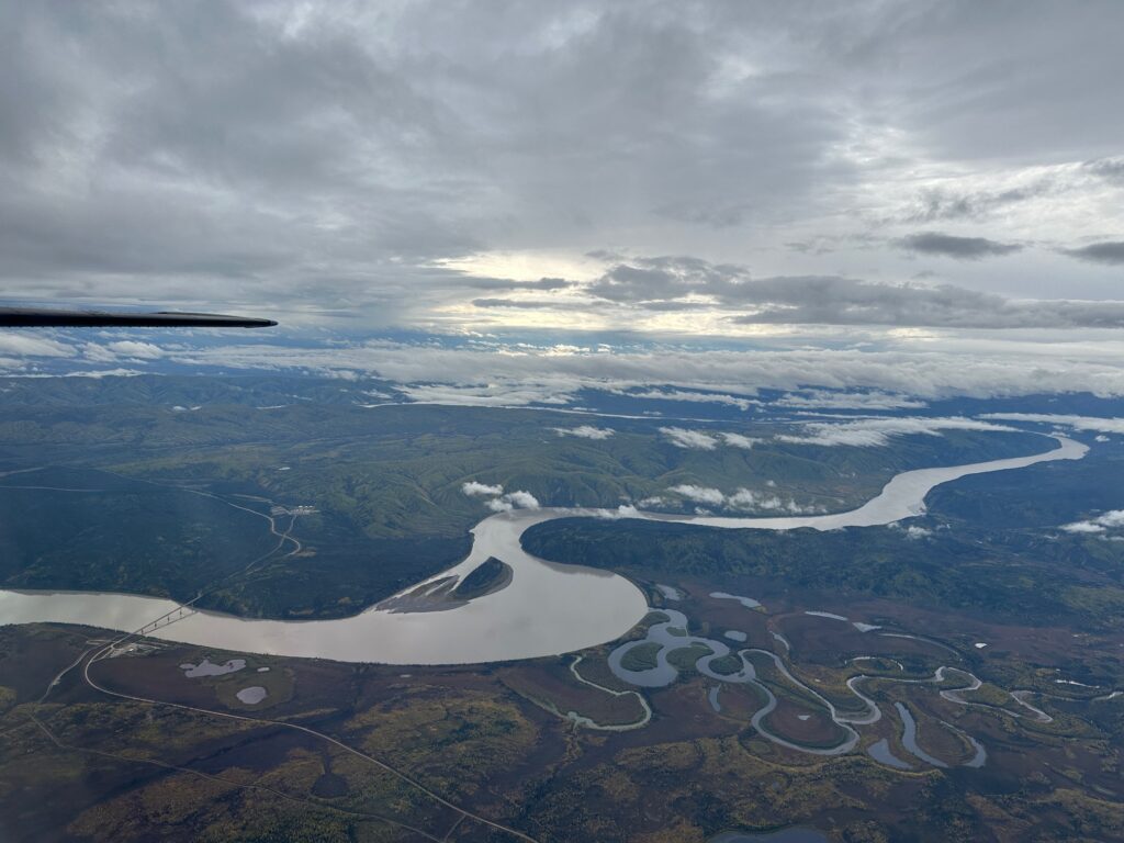 Aerial view of the Yukon River snaking through lush green hills and valleys under a cloudy sky, with the Yukon River Bridge visible below.